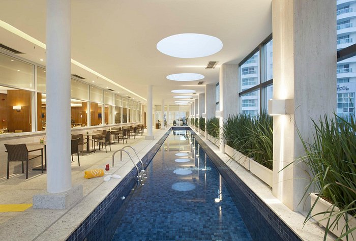 Foto da área interna de um hotel da Rede Windsor em Brasília. Na foto, uma piscina interna com plantas na lateral com mesas.