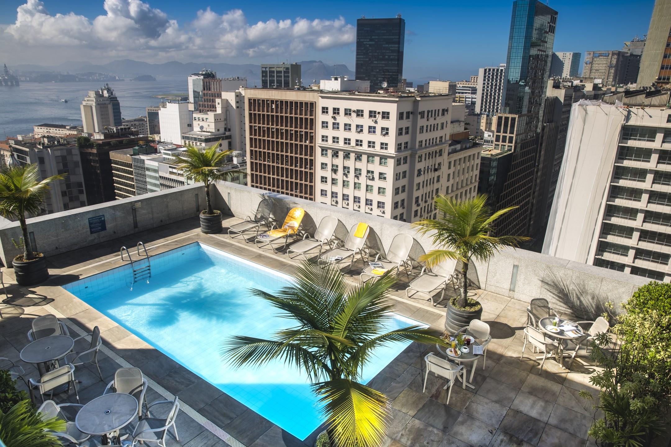 Foto da cobertura de um hotel da Rede Windsor no Centro do Rio. Na foto, há uma piscina, plantas e espreguiçadeiras. Ao redor, prédios do Centro do Rio.