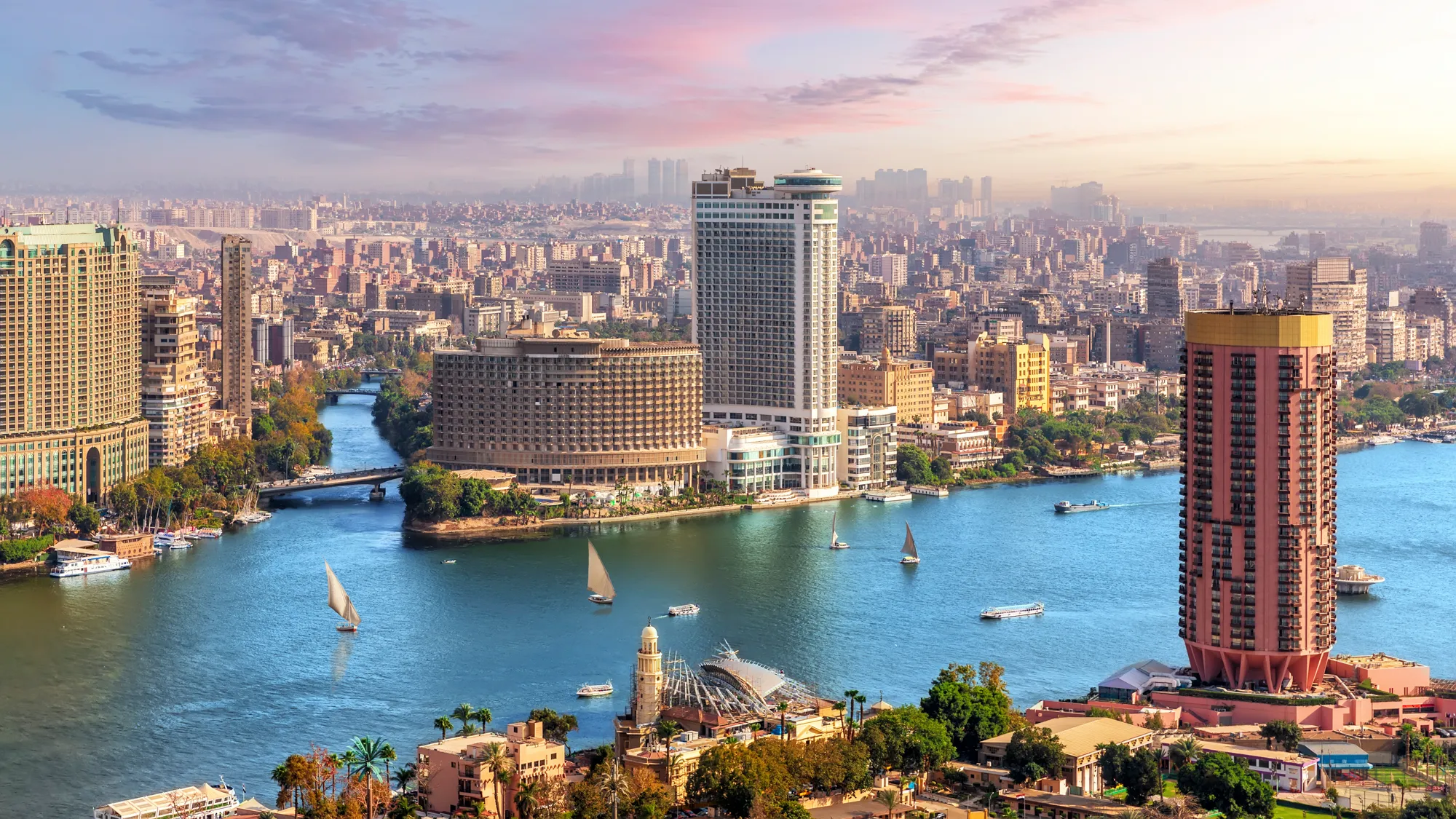 A imagem mostra uma vista panorâmica da cidade do Cairo, com destaque para o rio Nilo fluindo através da cidade. Há várias embarcações, incluindo barcos à vela tradicionais, navegando no rio. À beira do rio, observam-se diversos edifícios altos e modernos, misturados com arquitetura mais tradicional, indicando uma fusão de estilos antigos e contemporâneos. O céu apresenta tons suaves de azul e rosa, sugerindo que a imagem foi tirada próximo ao pôr ou nascer do sol. Esta vista da cidade transmite uma sensação de vivacidade e a mistura única de história e modernidade que Cairo oferece.