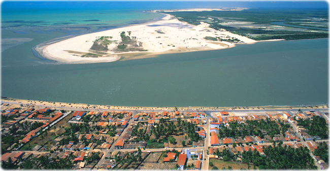 imagem aérea da cidade de Camocim, que consiste em uma área com casas e árvores, uma faixa de areia, mar e uma ilha