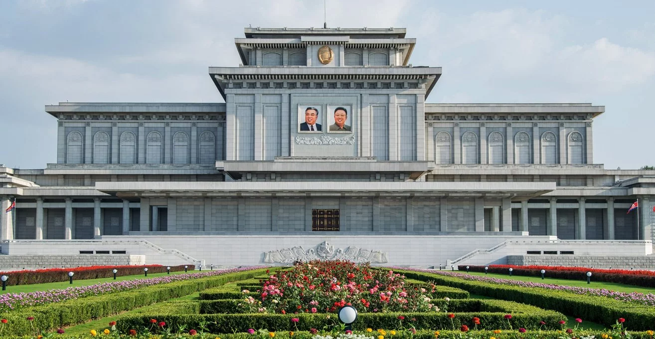 Vista frontal do Palácio do Sol de Kumsusan em Pyongyang, destacando sua arquitetura grandiosa com uma fachada cinza e adornos dourados sob um céu claro. No centro, imagens dos líderes norte-coreanos Kim Il-sung e Kim Jong-il são exibidas acima de um jardim simetricamente organizado com flores e arbustos bem cuidados.