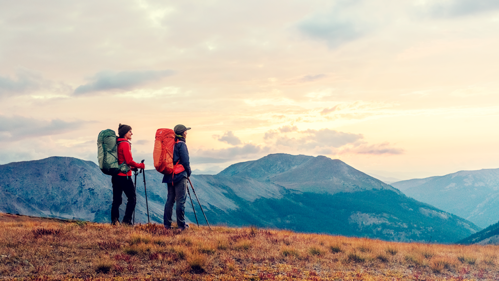 Dois aventureiros de costas observando a paisagem montanhosa ao entardecer, com mochilas grandes, prontos para explorar.