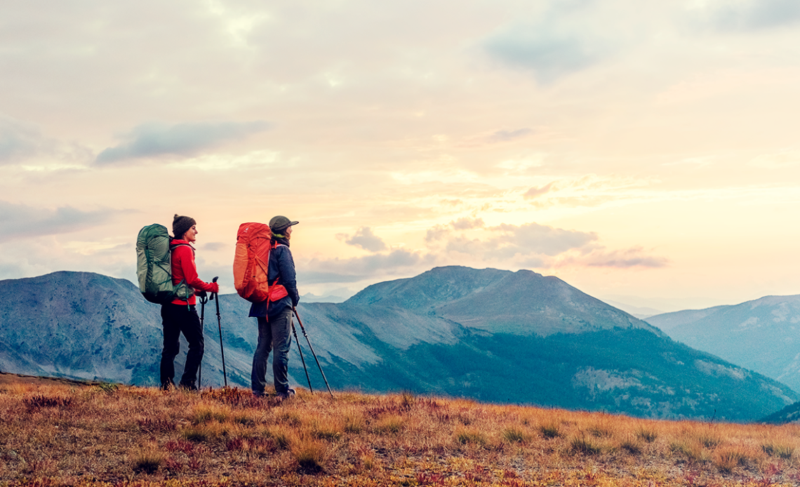 Dois aventureiros de costas observando a paisagem montanhosa ao entardecer, com mochilas grandes, prontos para explorar.