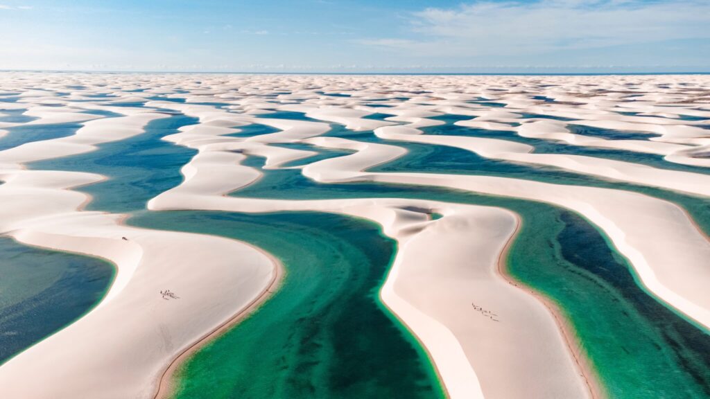Vista aérea dos Lençóis Maranhenses, mostrando dunas de areia branca intercaladas com lagoas de água azul turquesa, sob um céu claro.