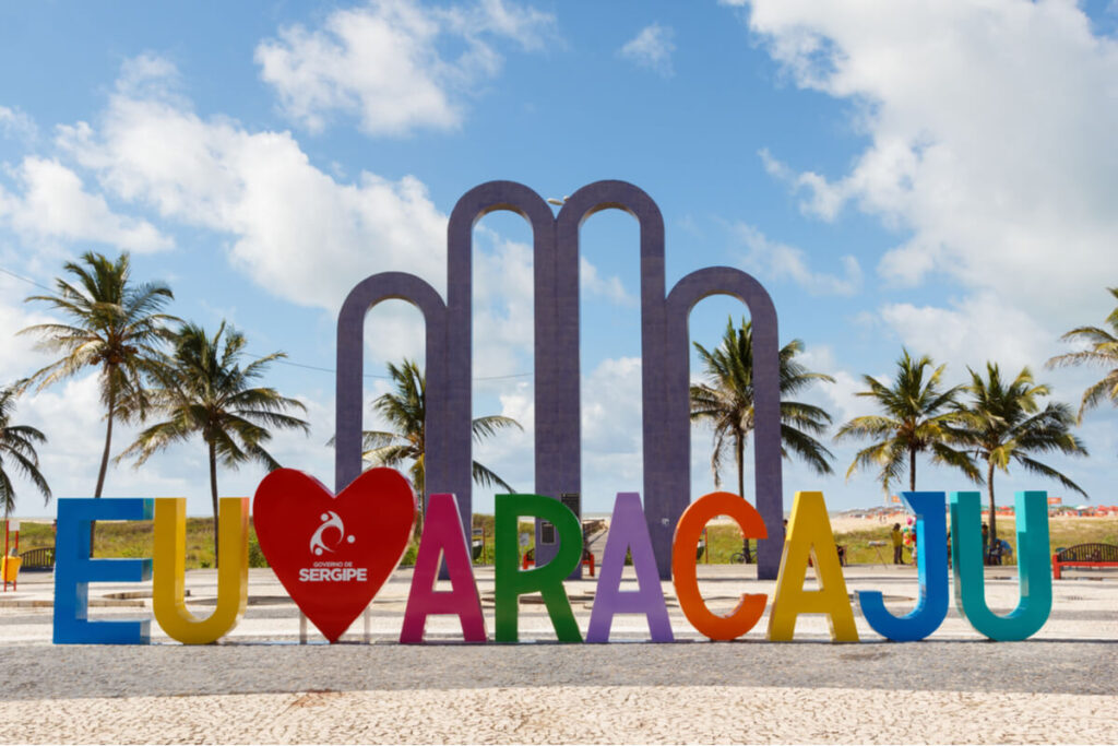 Letreiro colorido 'Eu ❤️ Aracaju' na Praia de Atalaia, em Aracaju, com céu azul, nuvens e palmeiras ao fundo.