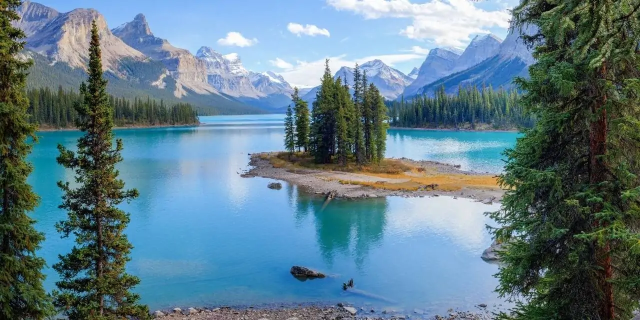 A imagem mostra uma paisagem natural com um lago de águas claras e cor azul-turquesa. No centro do lago, há uma pequena ilha com pinheiros. Ao fundo, montanhas com picos cobertos de neve sob um céu azul com poucas nuvens. Ao redor do lago, há uma floresta de árvores.