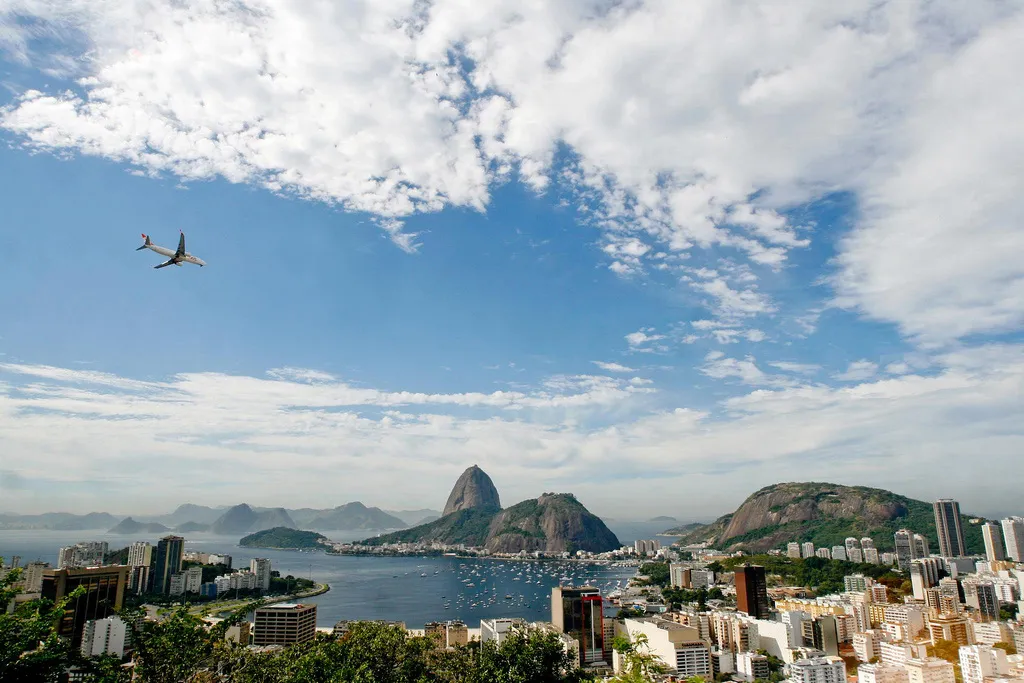 Vista aérea do Rio de Janeiro com um avião voando sobre a cidade e o Pão de Açúcar ao fundo.