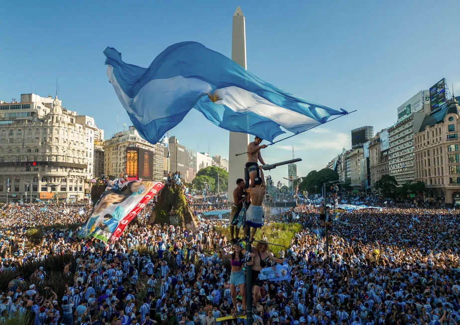 Multidão de fãs argentinos celebrando com uma grande bandeira da Argentina em frente ao Obelisco de Buenos Aires.