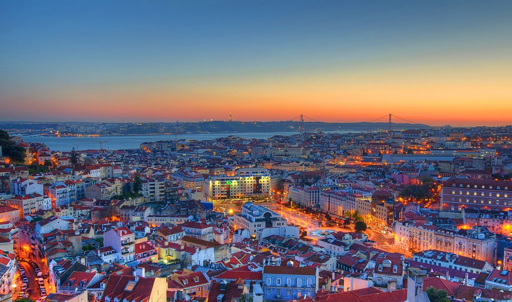 Vista panorâmica de Lisboa, Portugal, ao entardecer, mostrando telhas vermelhas, o rio Tejo e a Ponte 25 de Abril, com a cidade iluminada.