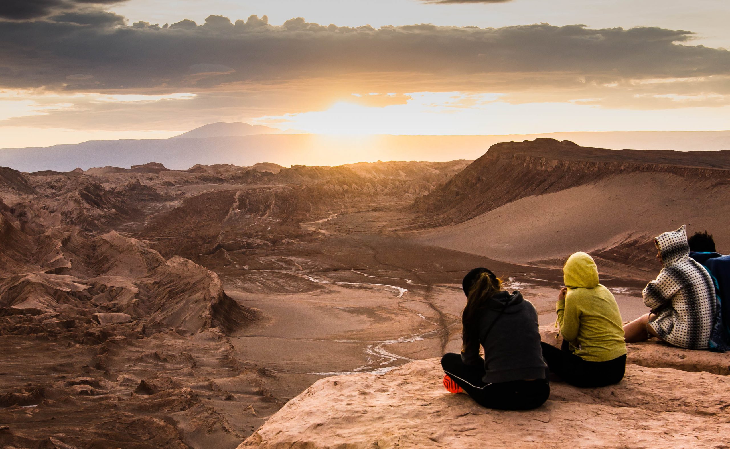 Grupo de amigos sentados no alto de uma colina, admirando o pôr do sol sobre o deserto de San Pedro de Atacama.