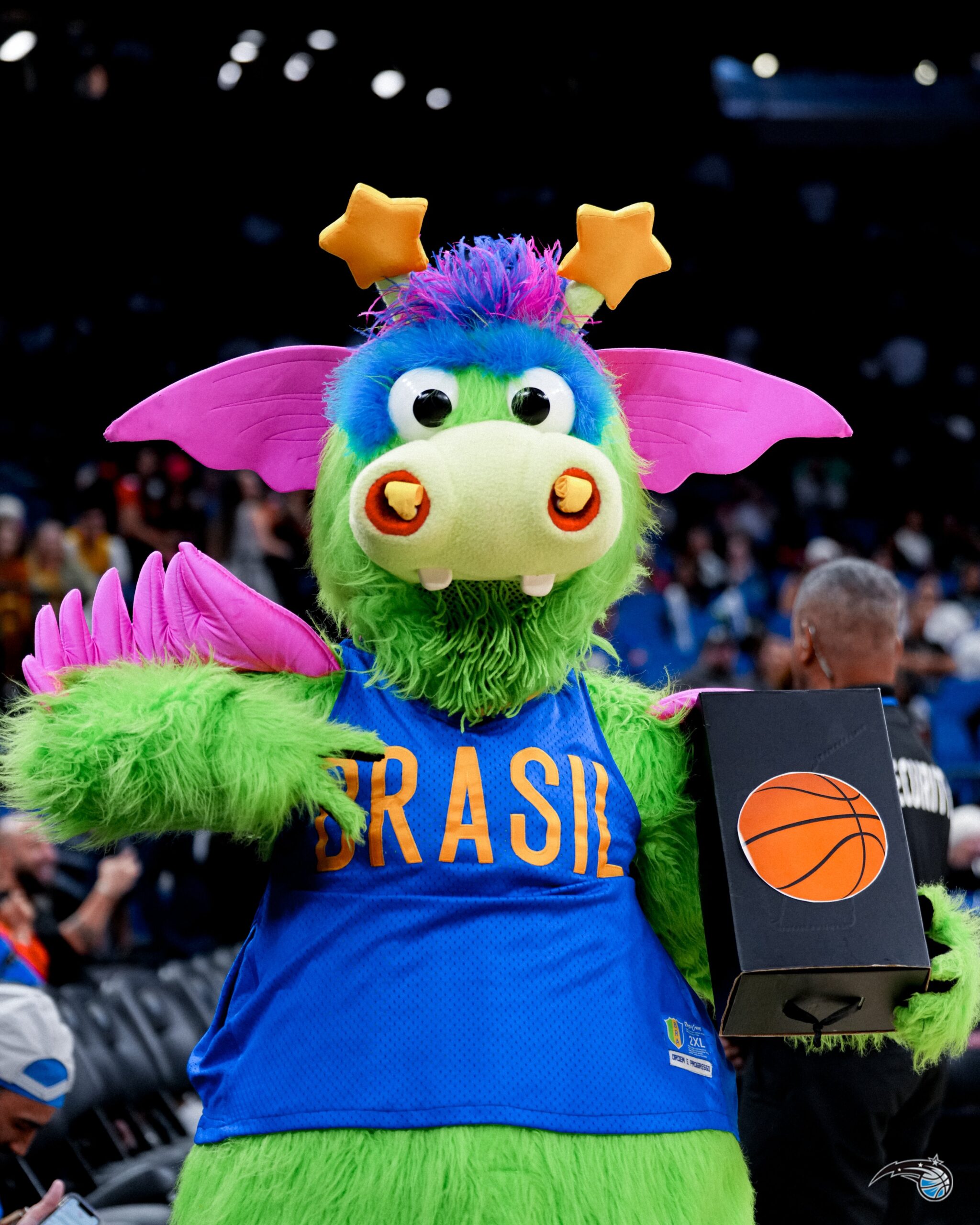 Stuff, a mascote do Orlando Magic, vestindo uma camisa do Brasil e segurando uma caixa com o símbolo de uma bola de basquete durante um evento de basquete.