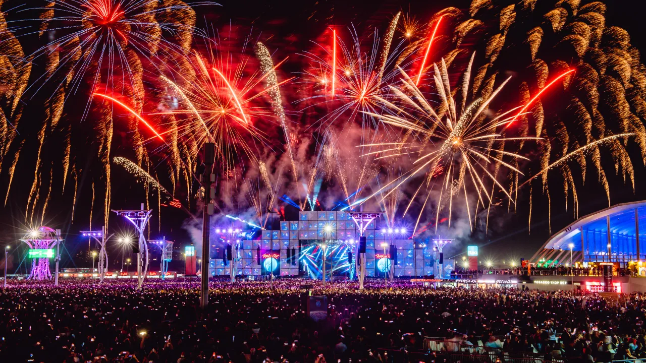 Fogos de artifício iluminam o céu noturno sobre o palco principal do Rock in Rio, com uma multidão de espectadores apreciando o espetáculo.