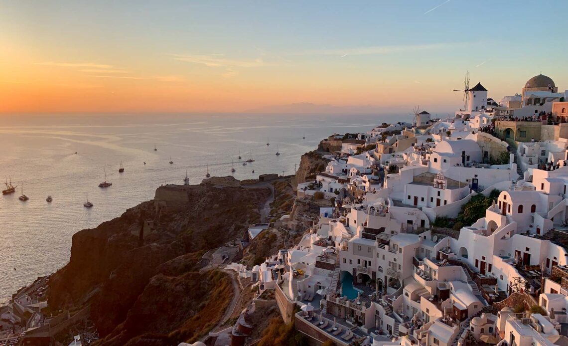 Vista de Oia, Santorini, ao pôr do sol, mostrando casas brancas, moinhos de vento e barcos no mar Egeu, iluminados pela luz dourada do entardecer.