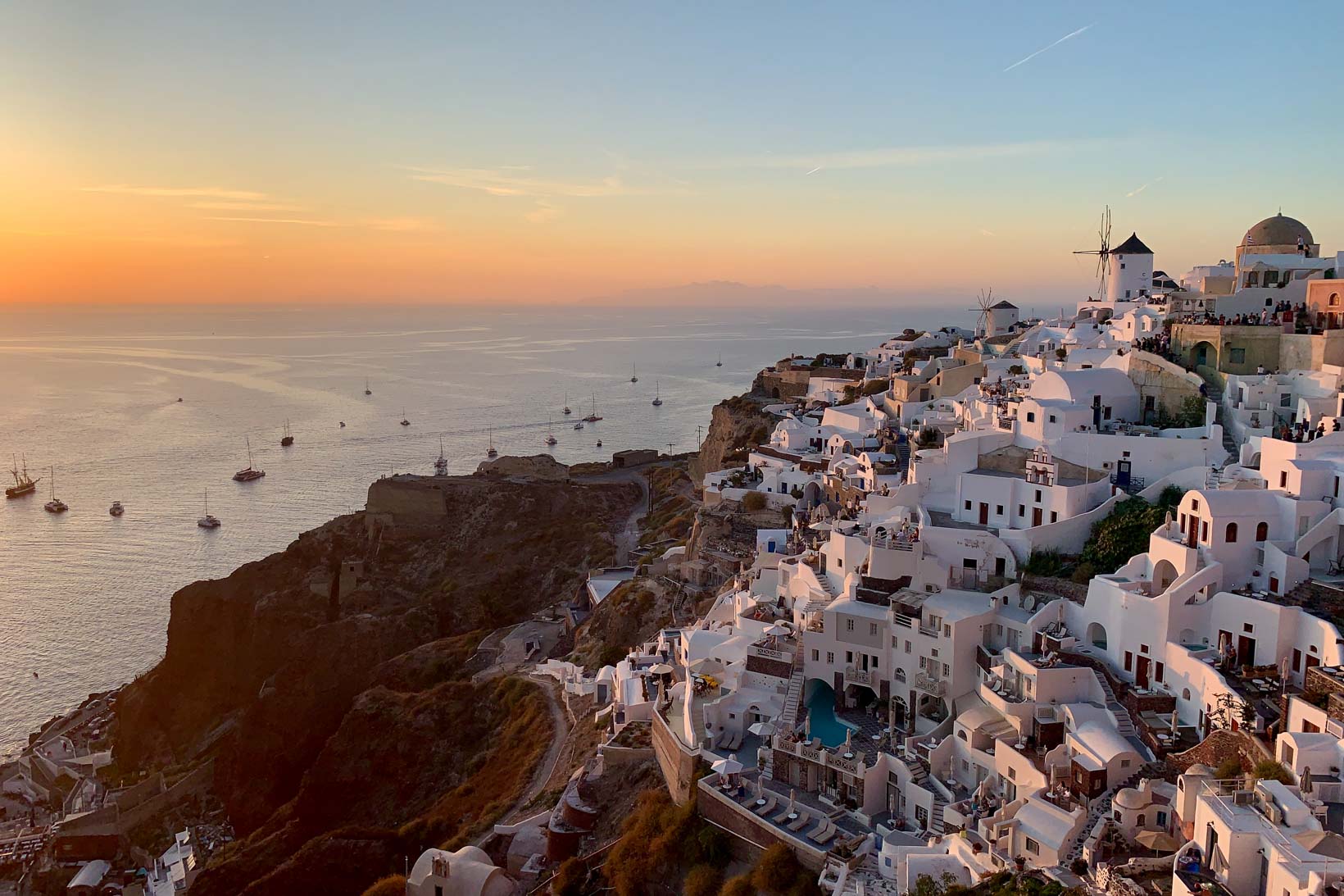 Vista de Oia, Santorini, ao pôr do sol, mostrando casas brancas, moinhos de vento e barcos no mar Egeu, iluminados pela luz dourada do entardecer.