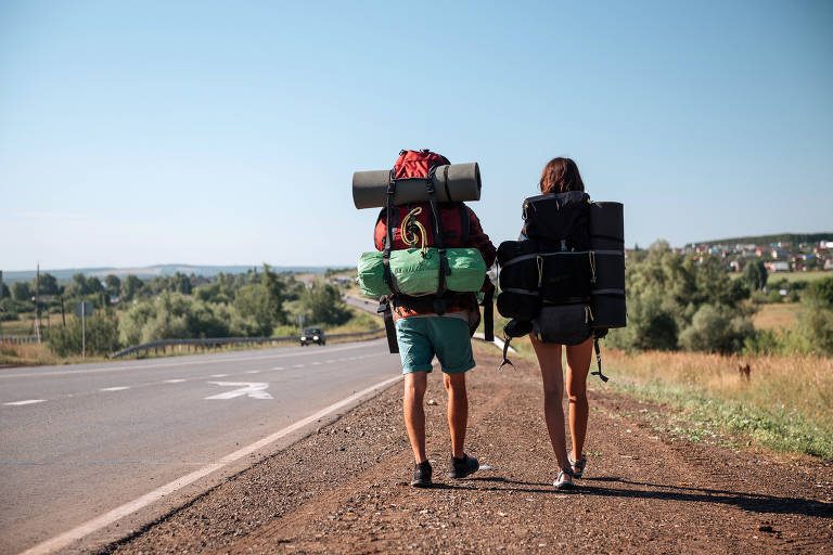 Duas pessoas de mochilas grandes caminhando ao lado de uma estrada rural.