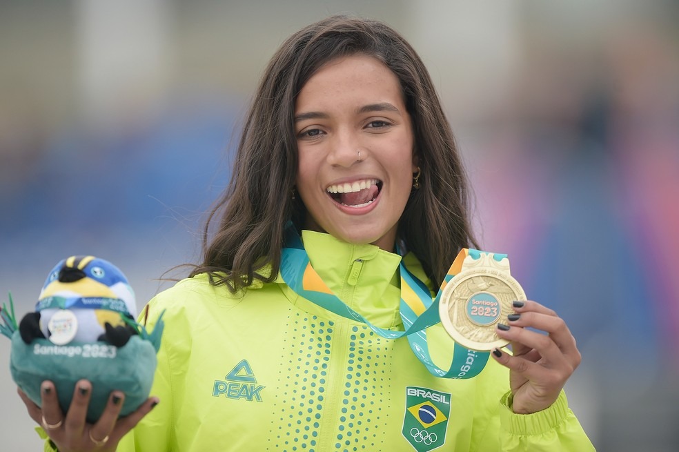 Rayssa Leal sorrindo, segurando uma medalha de ouro e um mascote dos Jogos Pan-Americanos Santiago 2023.