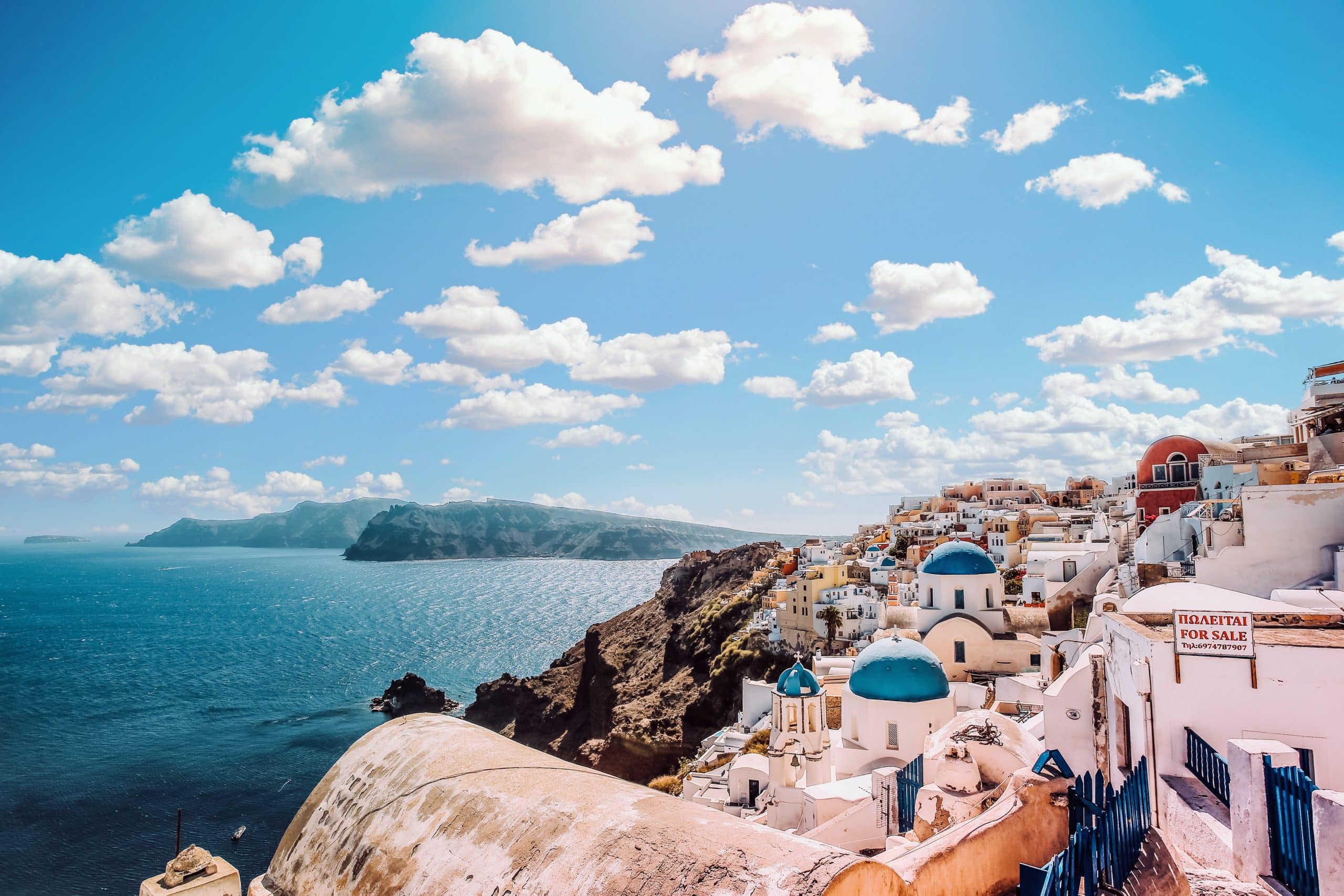 Imagem de Oia, Santorini, com três cúpulas azuis e casas brancas tradicionais, além de uma bandeira grega ao fundo, em um cenário de céu azul.