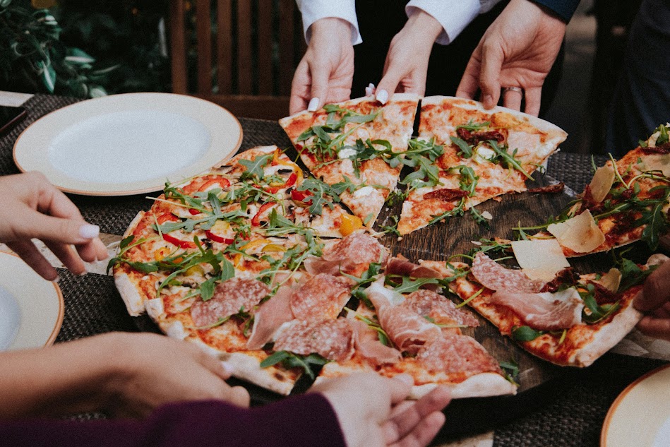 Várias pessoas pegando fatias de uma pizza grande, decorada com rúcula e fatias de presunto, em uma mesa ao ar livre.