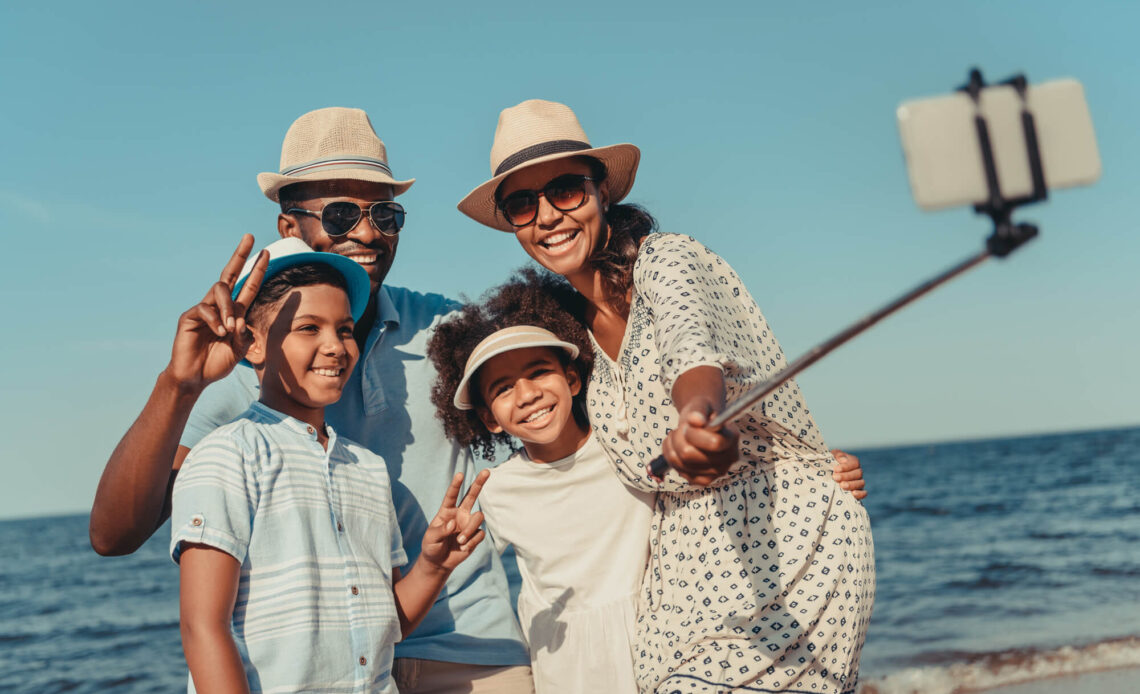 Família multigeracional sorrindo e tirando uma selfie na praia, representando o turismo multigeracional, uma tendência crescente em viagens.