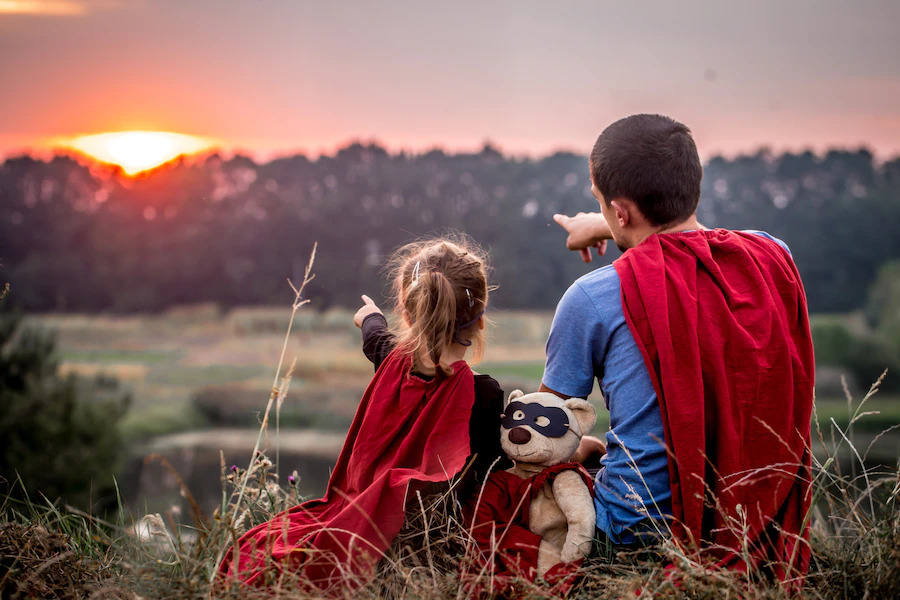 Pai e filha vestidos de super-heróis observando o pôr do sol durante uma viagem de Dia dos Pais.