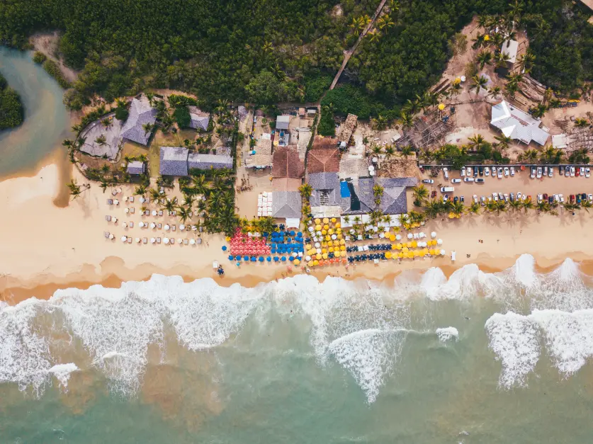 Vista aérea de Trancoso mostrando a Praia dos Nativos com guarda-sóis coloridos, construções rústicas e vegetação ao fundo.