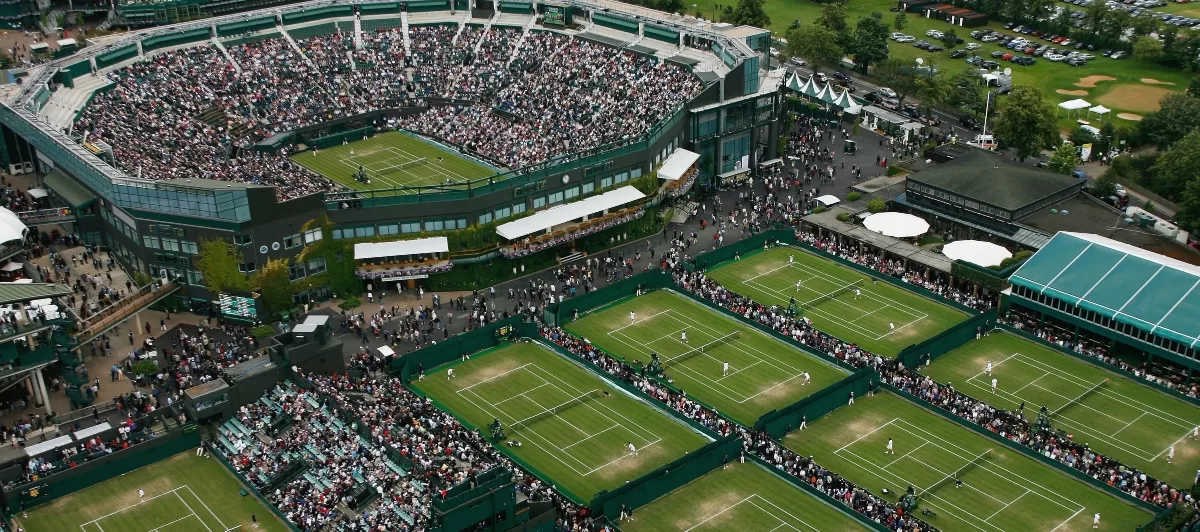 Vista aérea do estádio de Wimbledon, em Londres, durante um torneio de tênis, com quadras cheias de espectadores.