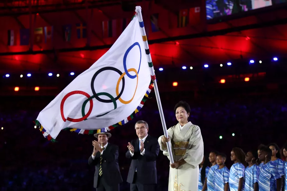 Uma mulher vestindo um kimono tradicional japonês segurando a bandeira olímpica durante a cerimônia de encerramento dos Jogos Olímpicos de Tóquio, com outras pessoas aplaudindo ao fundo.