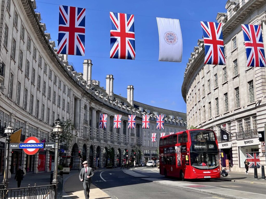 Rua de Londres decorada com bandeiras do Reino Unido, com um icônico ônibus vermelho de dois andares e uma placa do metrô em destaque.