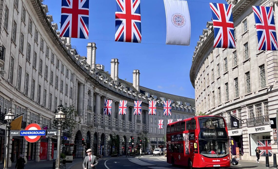 Rua de Londres decorada com bandeiras do Reino Unido, com um icônico ônibus vermelho de dois andares e uma placa do metrô em destaque.