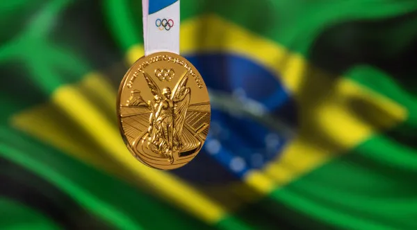 Medalha de ouro olímpica em destaque com a bandeira do Brasil ao fundo, representando o sucesso dos esportes brasileiros nas Olimpíadas. Medalha de ouro olímpica em primeiro plano, com a bandeira do Brasil desfocada ao fundo.