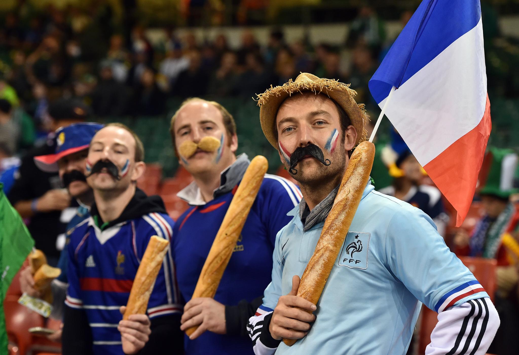 Torcedores franceses vestidos com fantasias e segurando baguetes, exibindo uma bandeira da França e bigodes falsos durante um evento esportivo.