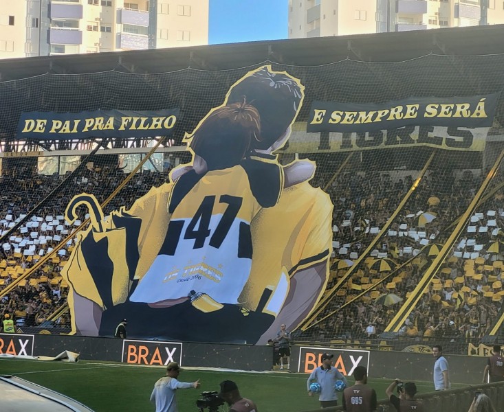 Pai e filho abraçados em frente a um mosaico no estádio, com a frase "De pai pra filho e sempre será"