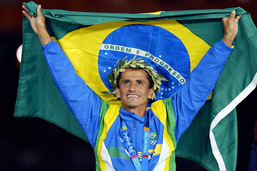 Atleta brasileiro segurando a bandeira do Brasil e usando uma coroa de louros durante a cerimônia de premiação nas Olimpíadas da cidade de Atenas.