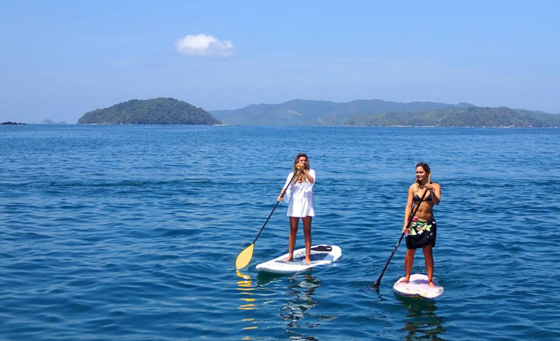 Duas pessoas praticando stand-up paddle em águas cristalinas, explorando destinos mostrados em conteúdos de televisão sobre aventuras globais.