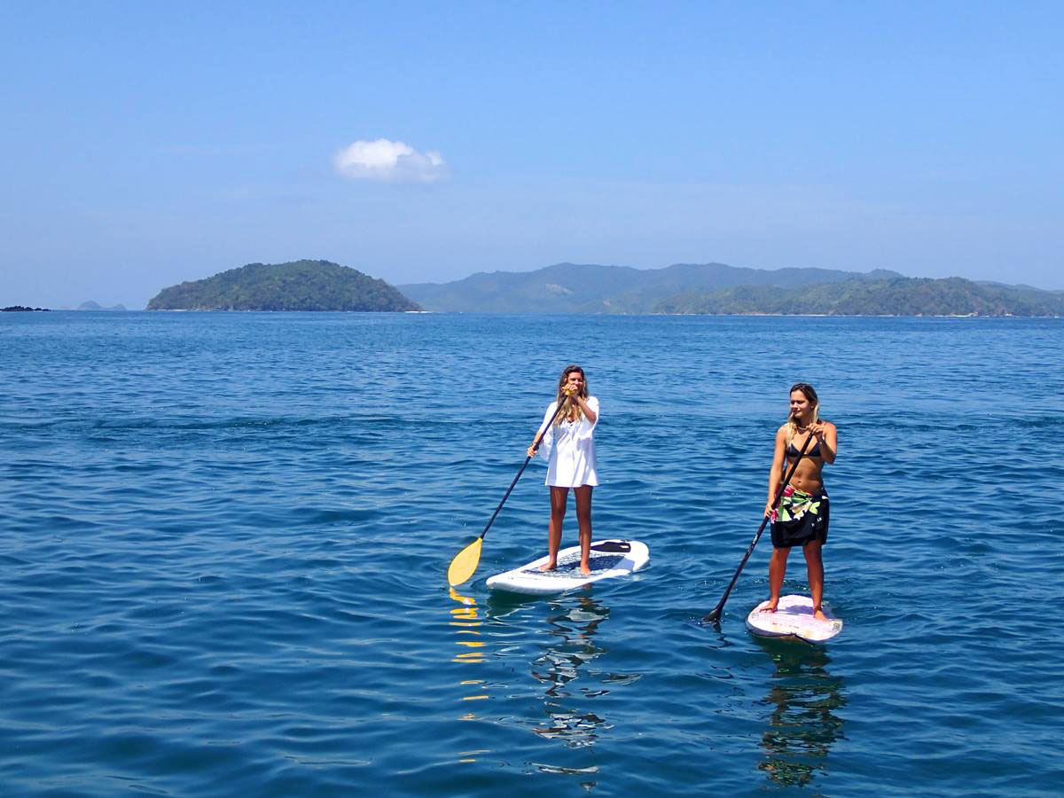 Duas pessoas praticando stand-up paddle em águas cristalinas, explorando destinos mostrados em conteúdos de televisão sobre aventuras globais.