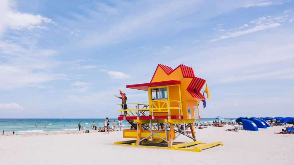 Posto de salva-vidas colorido em uma praia de Miami, Flórida, com banhistas relaxando na areia e o mar azul ao fundo.