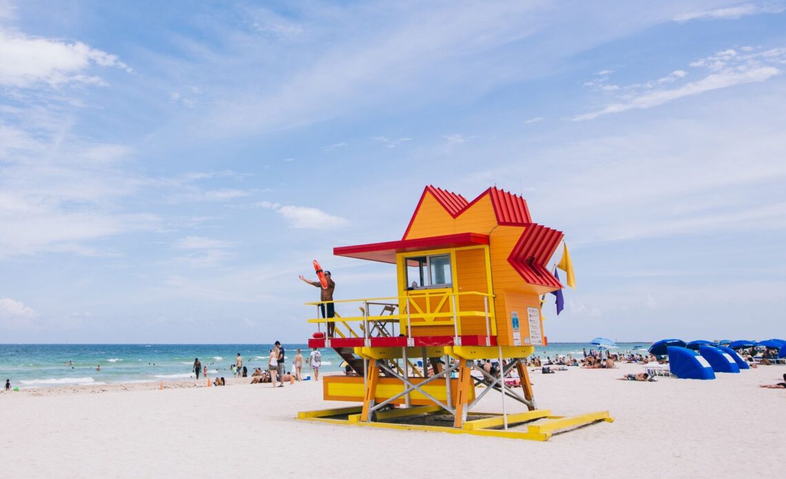 Posto de salva-vidas colorido em uma praia de Miami, Flórida, com banhistas relaxando na areia e o mar azul ao fundo.