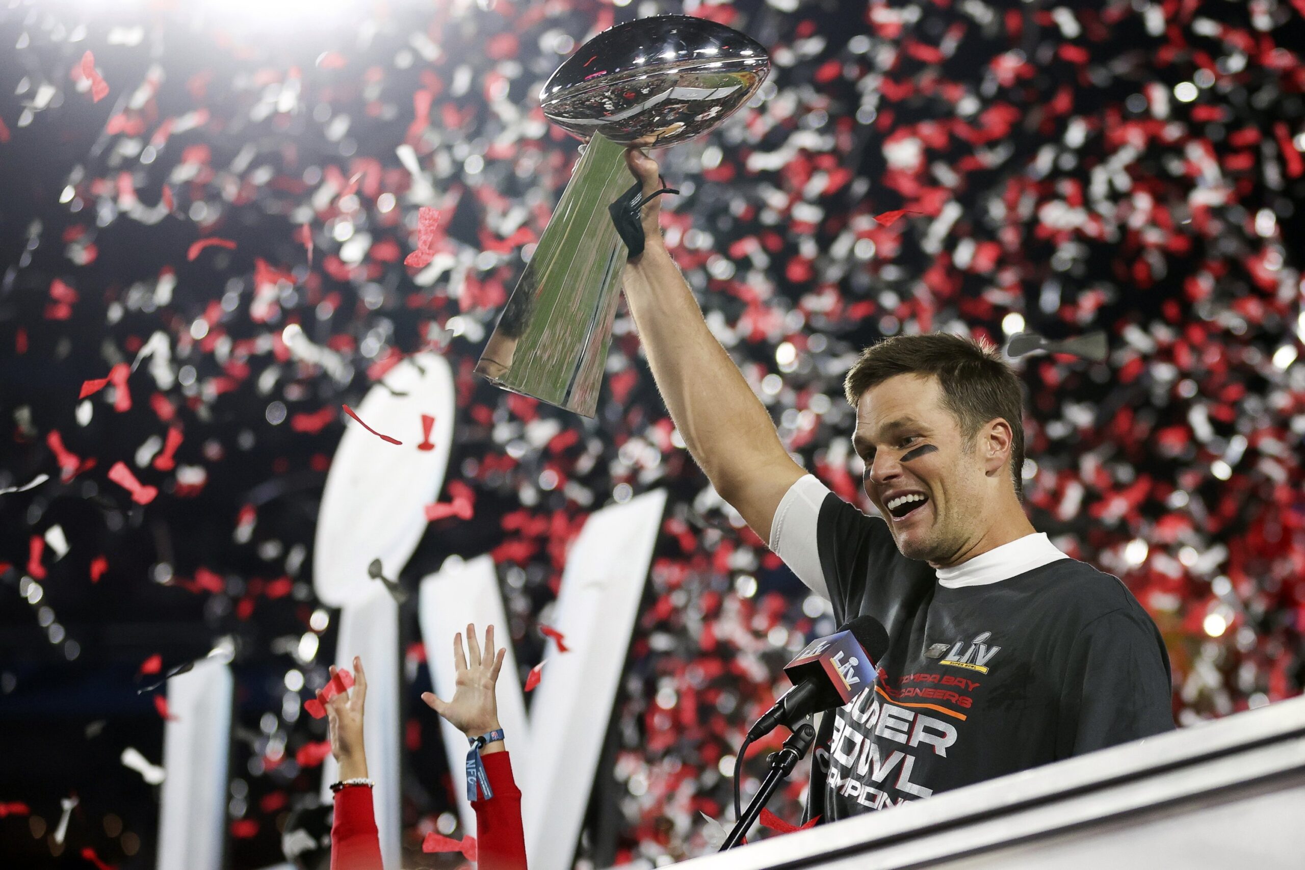 Tom Brady levantando o troféu do Super Bowl LIV com confetes vermelhos e brancos, cores do Tampa Bay Buccaneers, caindo em celebração.