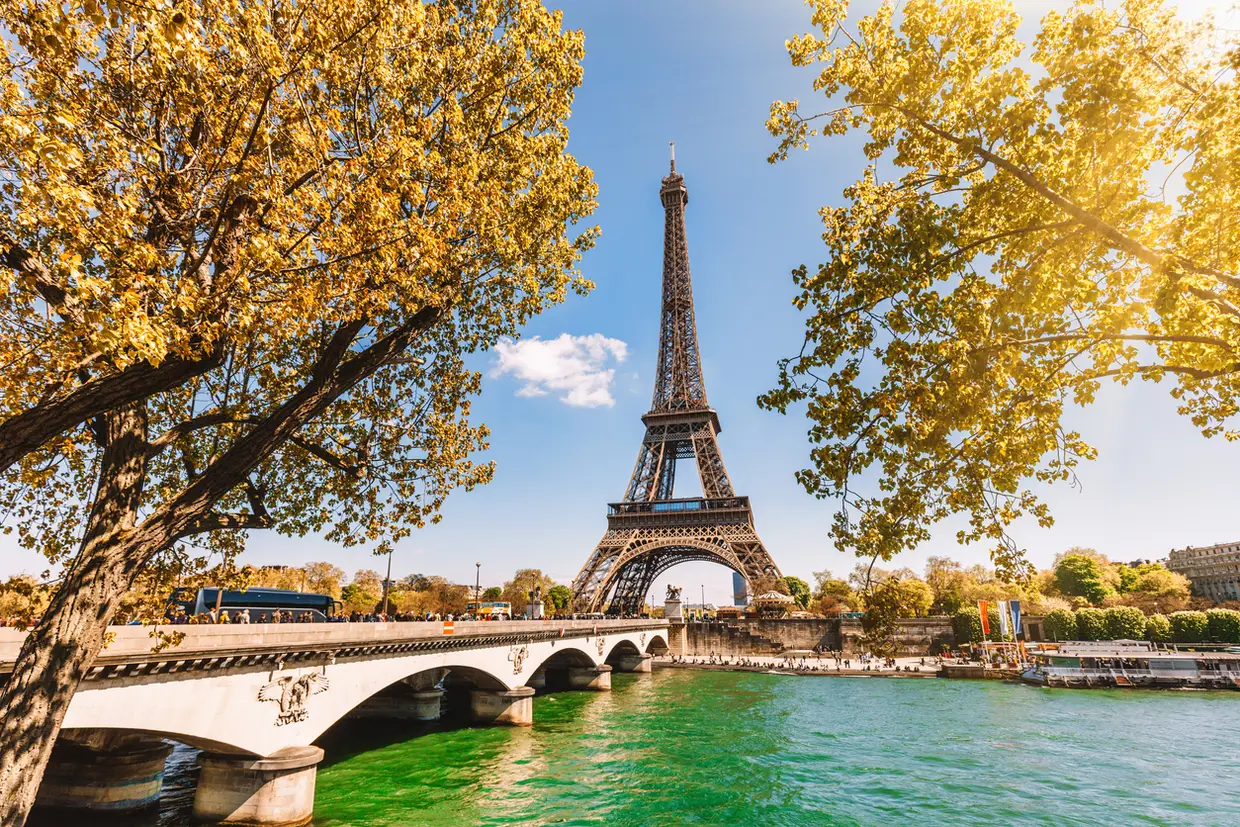 Vista da Torre Eiffel em Paris, com árvores douradas ao redor e o rio Sena ao fundo, em um dia ensolarado.