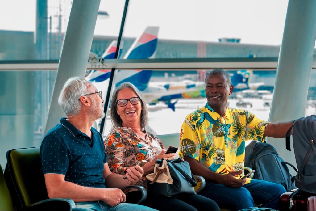 Grupo de aposentados sorrindo enquanto aguardam seu voo em um aeroporto, com aviões ao fundo. Voa Brasil.