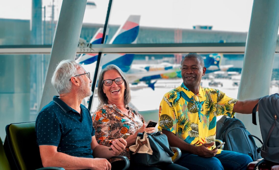 Grupo de aposentados sorrindo enquanto aguardam seu voo em um aeroporto, com aviões ao fundo. Voa Brasil.