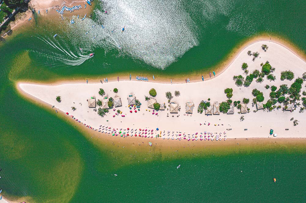 Vista aérea da Ilha do Amor em Alter do Chão, Pará, com águas verdes ao redor e uma praia de areia branca cheia de barracas e guarda-sóis coloridos.