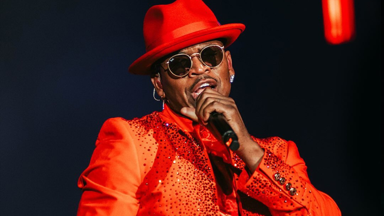 Ne-Yo cantando no Rock in Rio 2024, vestindo um elegante terno vermelho com chapéu combinando, segurando o microfone com estilo.