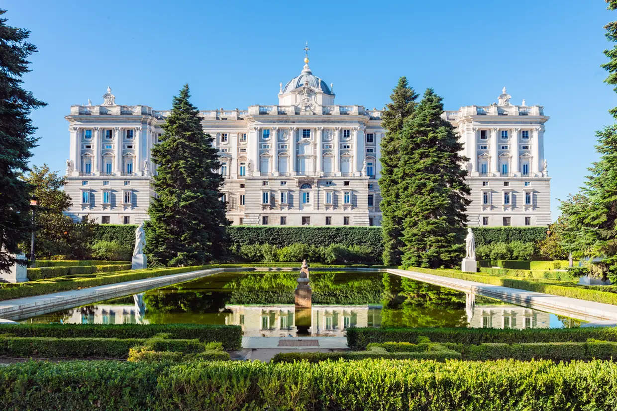 Palácio Real de Madrid visto dos jardins, com uma fonte refletindo o edifício em um dia claro e ensolarado.