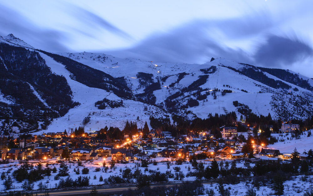 Vista noturna de Bariloche, Argentina, com luzes da cidade iluminando a base de montanhas cobertas de neve.