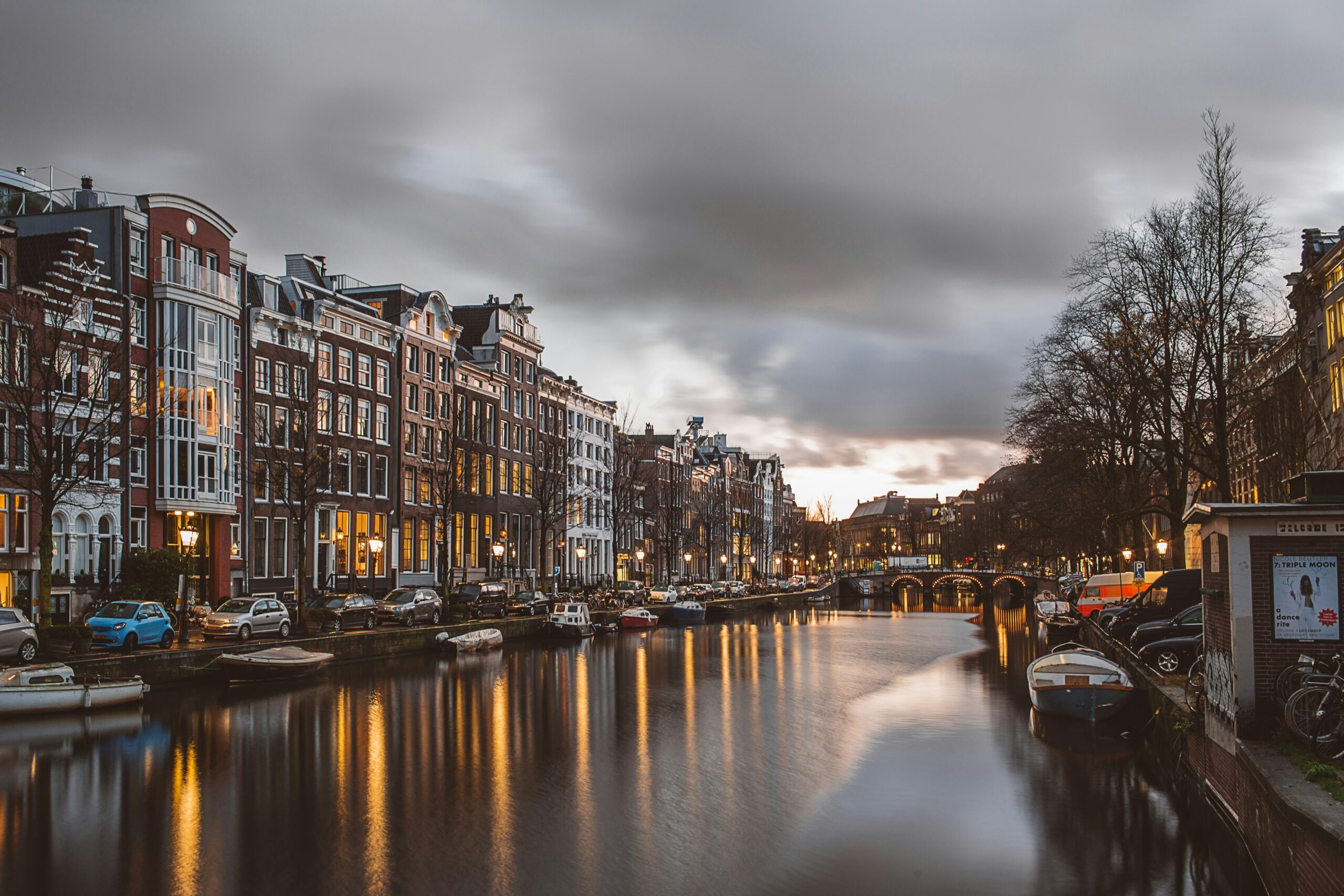 Canais de Amsterdã ao entardecer, refletindo as luzes dos prédios históricos e barcos ancorados, em um cenário típico da cidade.