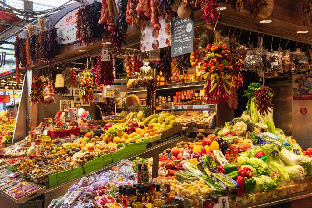 Barraca de frutas e vegetais em mercado gastronômico, com variedade de produtos frescos, incluindo pimentas, frutas e legumes.