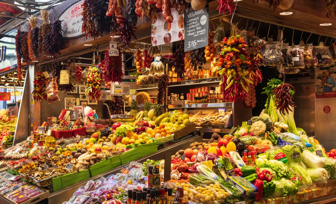 Barraca de frutas e vegetais em mercado gastronômico, com variedade de produtos frescos, incluindo pimentas, frutas e legumes.