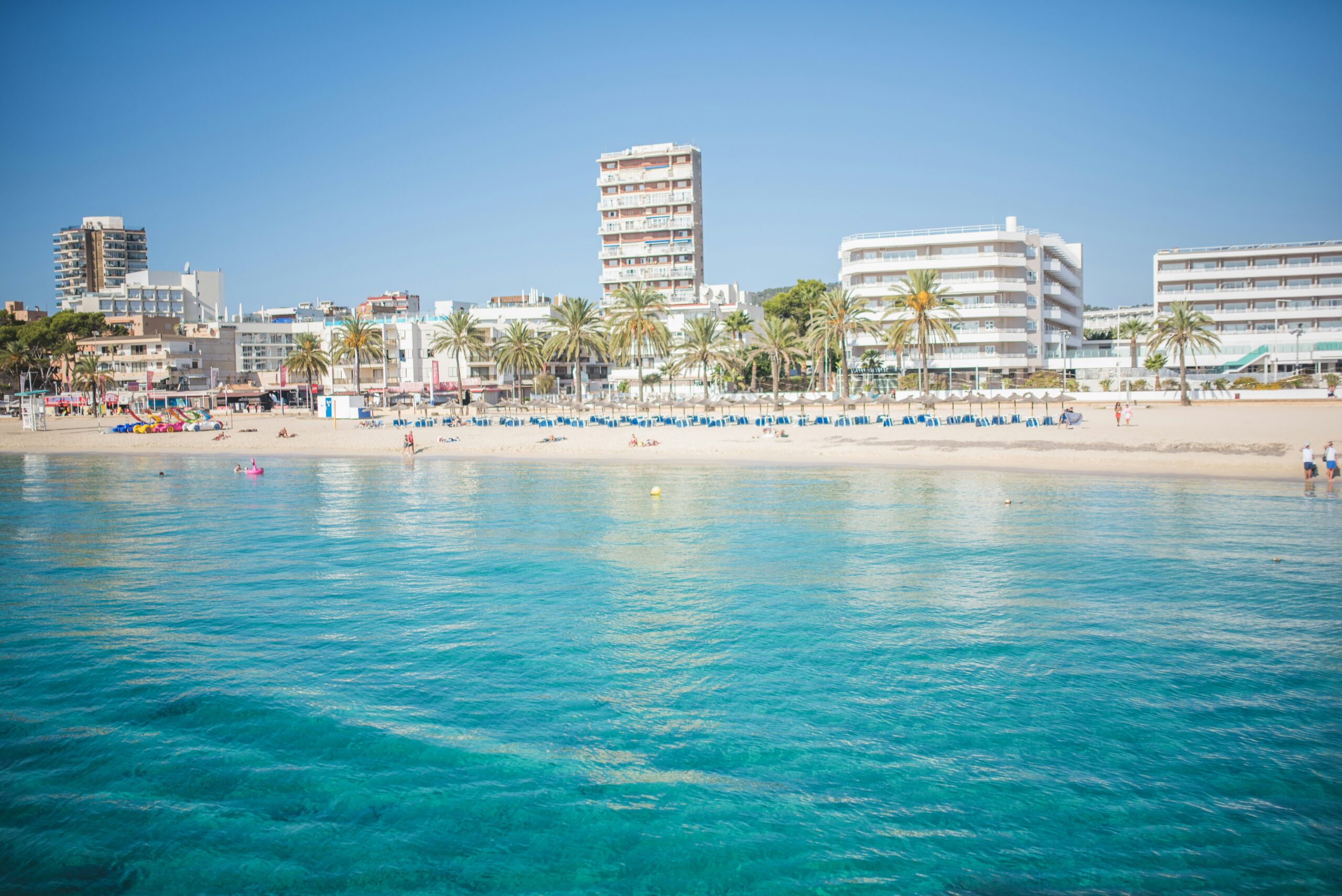 Praia em Mallorca, Espanha, com águas cristalinas e edifícios ao fundo.