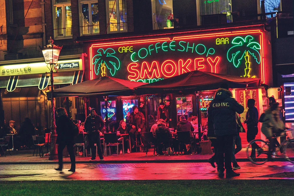 Fachada iluminada de um famoso coffeeshop em Amsterdã à noite, com bicicletas ao fundo e movimento de pessoas, caracterizando o turismo canábico da cidade.